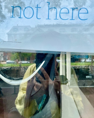 E reflected in glass shop window under sign that says, “not here”