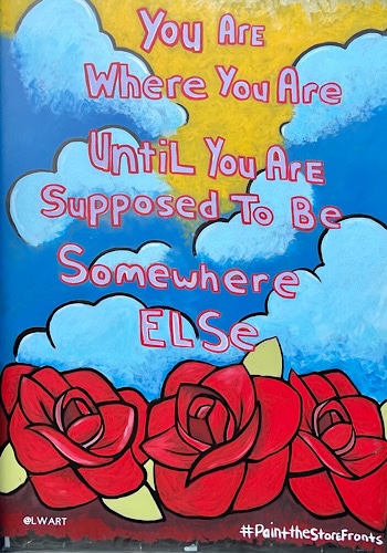 Painted storefront on background of sky and red roses, reads “you are where you are until you are supposed to be somewhere else.”
