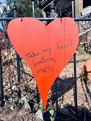 Heart-shaped red sign hung from wrought iron fence, with handwritten note:  “take my heart beating madly”
