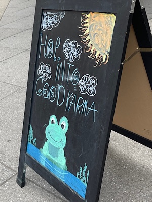 Sign outside a coffee shop has a drawing in chalk of a smiling from and reads, “hop into good karma.”