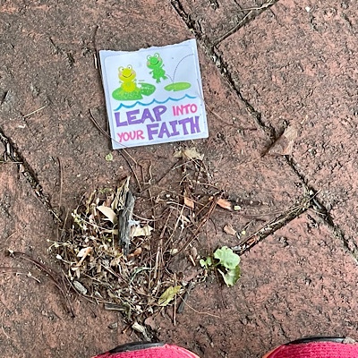 Photo of a discarded sticker on a brick sidewalk, with the tip of E’s sneakers showing.  Sticker has cartoon images of smiling frogs and reads “jump into your faith.”