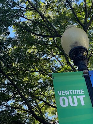 Sign attached to street light reads, “venture out.”