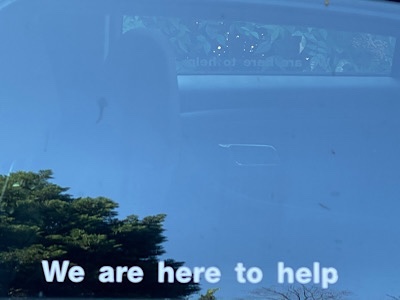 Photo of stenciled words on a window reflecting the sky and a tree that reads, “we are here to help.”