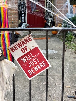 Halloween joke sign attached to fence in front of row house reads, “Be aware of/well just beware…”