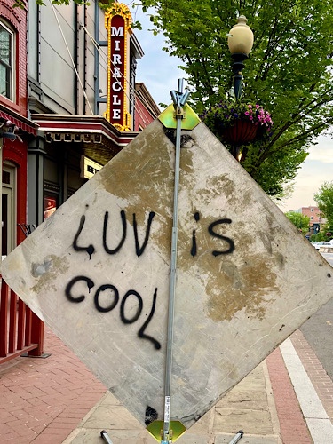 Graffiti that says “luv is cool” on back of street construction sign in front of marquee that says “miracle”
