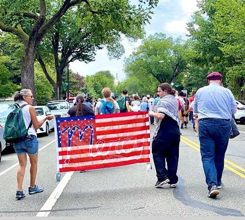 Photo of people marching to the United States Capitol holding a banner like an US Flag with the word “mercy” written on it.