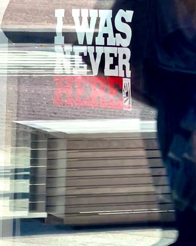 Sign viewed through window with the reflection of a person walking off frame, reads “I was never here.”