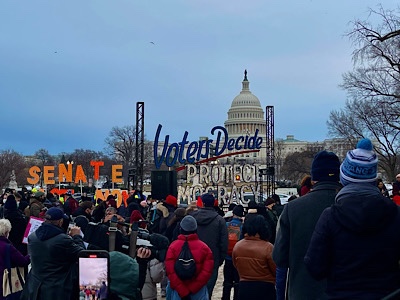 Image of demonstrators in front of United States Capitol Dome, with signs reading, “senate, act now” and “voters decide;protect democracy.”
