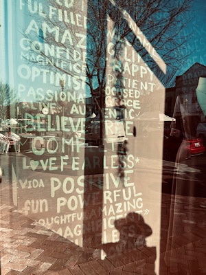 Image of shadow of photographer looking into window behind which is a wall of positive affirmations (to help sell clothing)